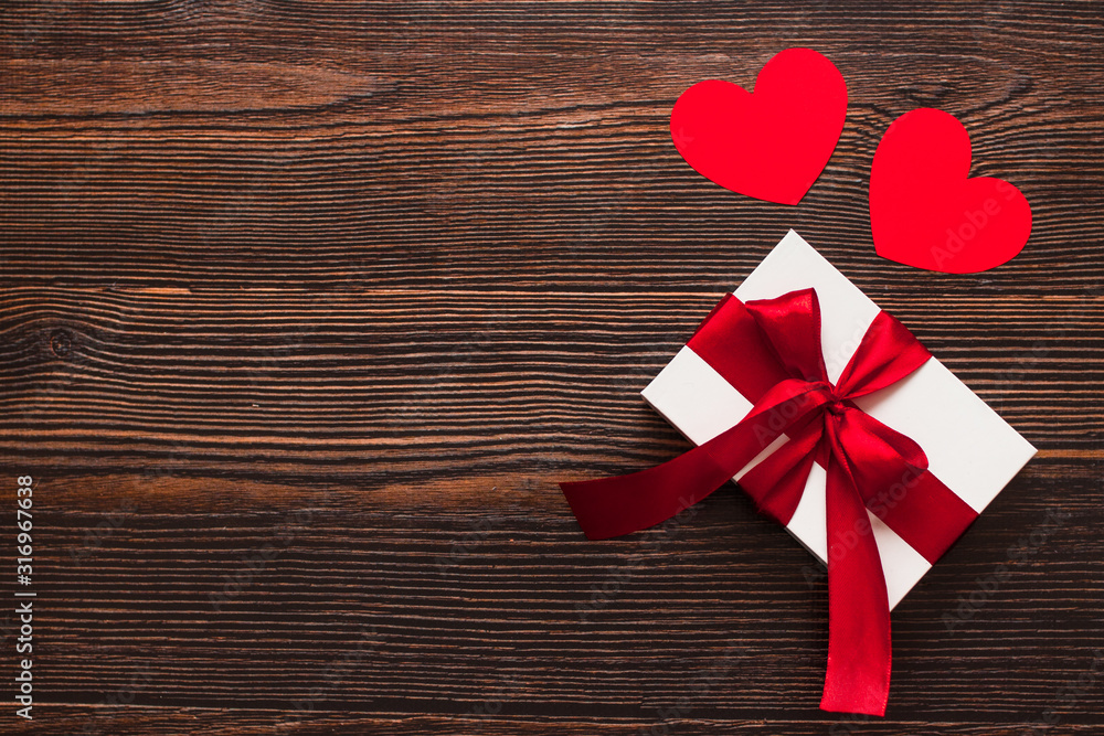 White present with a red ribbon and paper hearts isolated on a dark wooden background. Top side view of a celebrating warm flatlay. Valentines day and Christmas concept. Copyspace. White present with a red ribbon and paper hearts isolated on a dark wooden background. Top side view of a celebrating warm flatlay. Valentines day and Christmas concept. Copyspace.