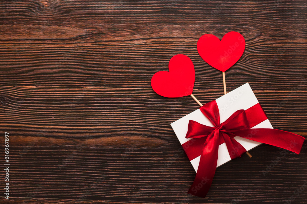 White present with a red ribbon and paper hearts isolated on a dark wooden background. Top side view of a celebrating warm flatlay. Valentines day and Christmas concept. Copyspace. White present with a red ribbon and paper hearts isolated on a dark wooden background. Top side view of a celebrating warm flatlay. Valentines day and Christmas concept. Copyspace.