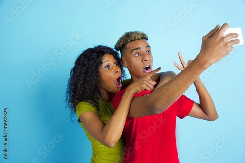 Taking selfie or vlog together. Young emotional african-american man and woman in colorful clothes on blue background. Beautiful couple. Concept of human emotions, facial expession, relations, ad.