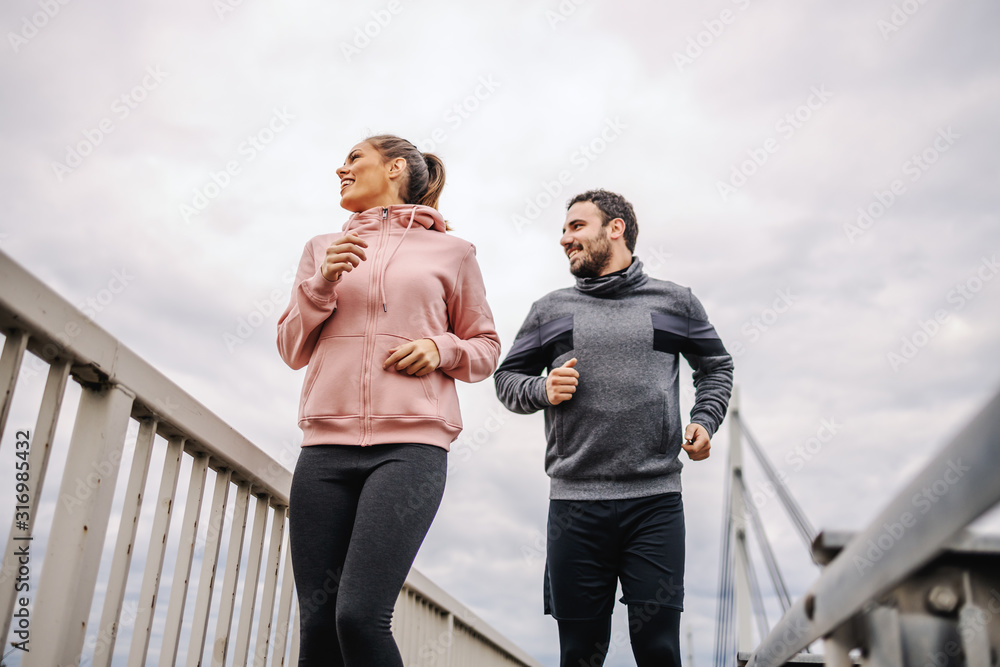 Low angle view of dedicated young heterosexual sporty friends in sportswear running on the bridge and looking at beautiful view. Urban exterior. Fitness outdoors concept.