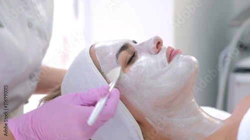 Cosmetologist is applying white mask on woman client face in beauty clinic. Portrait of woman, side view. Beautician making beauty facial skincare procedure to patient. Beauty industry concept.