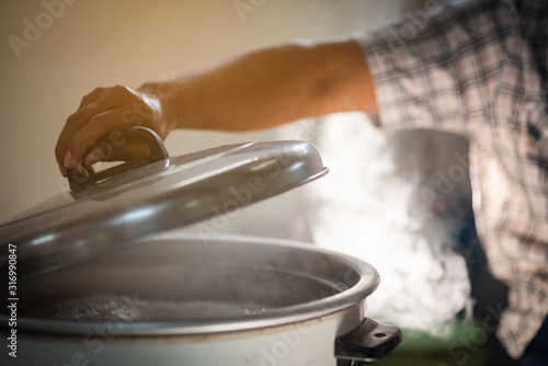 The chef is opening the lid of the rice cooker, the mass of steam reflected in the morning light coming out of a large electric rice cooker heated in the cafeteria.