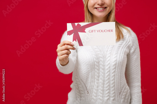Cropped image of young woman in white sweater hat isolated on red background studio portrait. Healthy fashion lifestyle people emotions, cold season concept. Mock up copy space. Hold gift certificate.