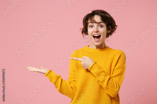 Cheerful excited young brunette woman girl in yellow sweater posing isolated on pastel pink background studio portrait. People lifestyle concept. Mock up copy space. Pointing index finger, hand aside. photo