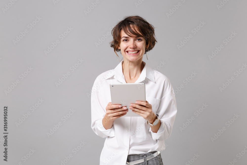 Smiling young business woman in white shirt posing isolated on grey wall background studio portrait. Achievement career wealth business concept. Mock up copy space. Holding, using tablet pc computer. Smiling young business woman in white shirt posing isolated on grey wall background studio portrait. Achievement career wealth business concept. Mock up copy space. Holding, using tablet pc computer.