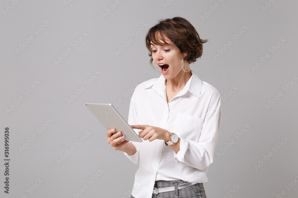 Shocked young business woman in white shirt posing isolated on grey wall background studio portrait. Achievement career wealth business concept. Mock up copy space. Holding, using tablet pc computer. Shocked young business woman in white shirt posing isolated on grey wall background studio portrait. Achievement career wealth business concept. Mock up copy space. Holding, using tablet pc computer.