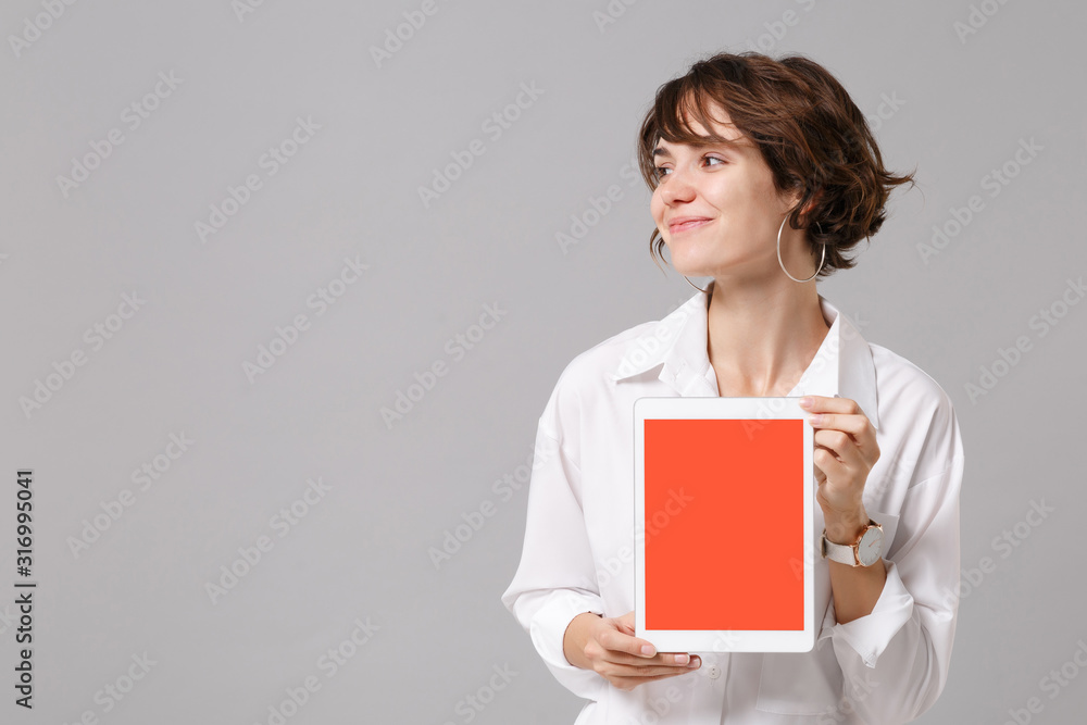 Pretty business woman in white shirt posing isolated on grey background. Achievement career wealth business concept. Mock up copy space. Hold tablet pc computer with blank empty screen, looking aside. Pretty business woman in white shirt posing isolated on grey background. Achievement career wealth business concept. Mock up copy space. Hold tablet pc computer with blank empty screen, looking aside.