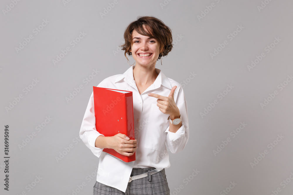 Smiling young business woman in white shirt posing isolated on grey background. Achievement career wealth business concept. Mock up copy space. Pointing index finger on red folder for papers document. Smiling young business woman in white shirt posing isolated on grey background. Achievement career wealth business concept. Mock up copy space. Pointing index finger on red folder for papers document.