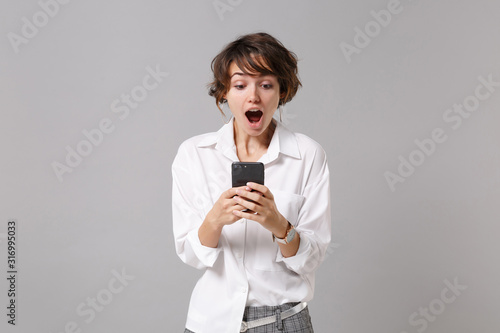 Shocked young business woman in white shirt posing isolated on grey background studio portrait. Achievement career wealth business concept. Mock up copy space. Using mobile phone, typing sms message. © ViDi Studio