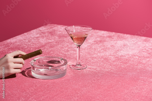 cropped view of woman holding cigar near ashtray and glass of rose wine on velour cloth isolated on pink, girlish concept