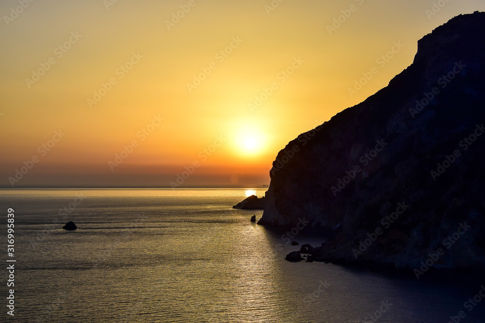 Paleokastritsa / Corfu, Greece - Sunset, yellow-orange sky, dark silhouette of a cliff, sea, in the summer afternoon.