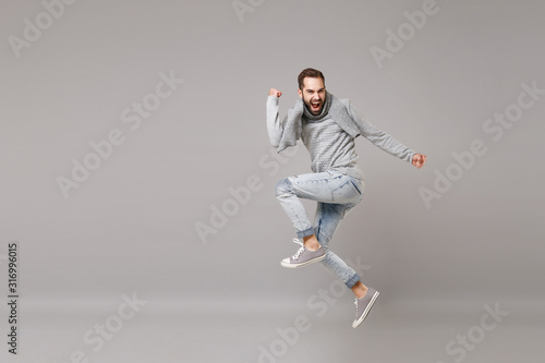 Young bearded man in gray sweater, scarf posing isolated on grey wall background, studio portrait. Healthy fashion lifestyle, cold season concept. Mock up copy space. Jumping, doing winner gesture.