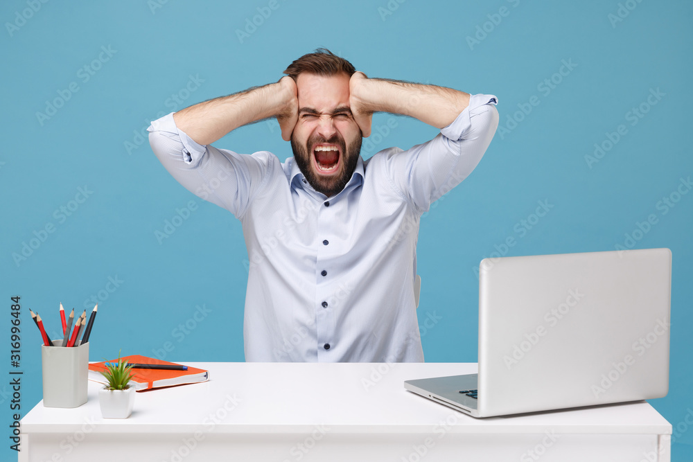 Frustrated man in shirt sit work at desk with pc laptop isolated on blue background. Achievement business career lifestyle concept. Mock up copy space. Screaming keeping eyes closed put hands on head. Frustrated man in shirt sit work at desk with pc laptop isolated on blue background. Achievement business career lifestyle concept. Mock up copy space. Screaming keeping eyes closed put hands on head.