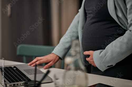 Close up of pregnant CEO standing in her office  touching belly and using laptop for checking an email from important client.