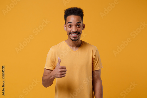 Smiling young african american guy in casual t-shirt posing isolated on yellow orange wall background, studio portrait. People sincere emotions lifestyle concept. Mock up copy space. Showing thumb up.