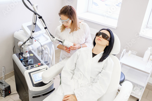 Charming female beautician does ultrasonic face cleaning pretty woman client sitting in a chair in a white coat and safety glasses in beauty spa clinic. ?oncept of skin cleansing procedures