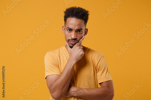 Preoccupied young african american guy in casual t-shirt posing isolated on yellow orange background studio portrait. People emotions lifestyle concept. Mock up copy space. Put hand prop up on chin. © ViDi Studio