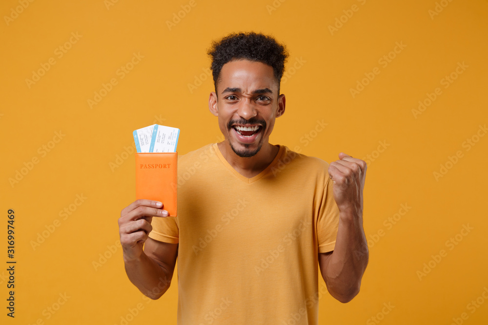 Happy young african american guy in casual t-shirt posing isolated on yellow orange background. People lifestyle concept. Mock up copy space. Hold passport boarding pass tickets, doing winner gesture. Happy young african american guy in casual t-shirt posing isolated on yellow orange background. People lifestyle concept. Mock up copy space. Hold passport boarding pass tickets, doing winner gesture.