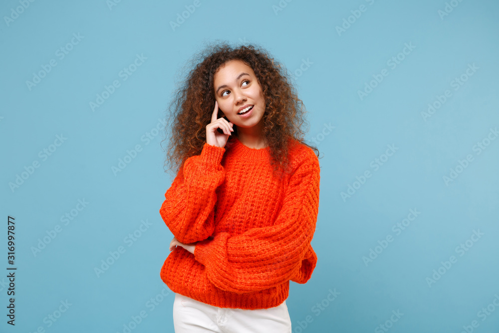 Smiling young african american girl in casual orange knitted clothes isolated on pastel blue background studio portrait. People sincere emotions, lifestyle concept. Mock up copy space. Looking aside. Smiling young african american girl in casual orange knitted clothes isolated on pastel blue background studio portrait. People sincere emotions, lifestyle concept. Mock up copy space. Looking aside.
