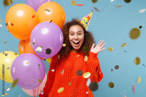 Amazed young african american girl in orange knitted clothes birthday hat isolated on pastel blue background. Holiday party concept. Celebrating with confetti, colorful air balloons spreading hands.