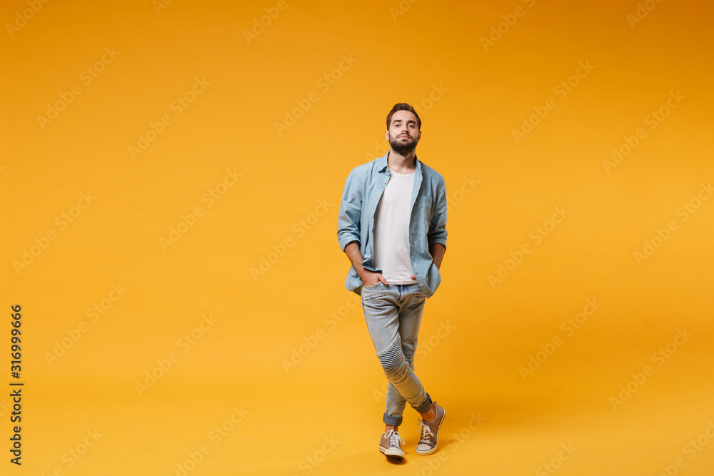 Handsome young bearded man in casual blue shirt posing isolated on yellow orange background, studio portrait. People sincere emotions lifestyle concept. Mock up copy space. Holding hands in pockets. - obrazy, fototapety, plakaty 