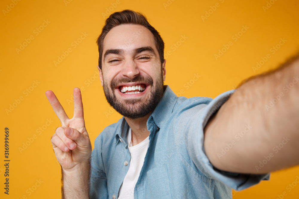 Close up of smiling young man in casual blue shirt posing isolated on yellow orange background. People lifestyle concept. Mock up copy space. Doing selfie shot on mobile phone showing victory sign.