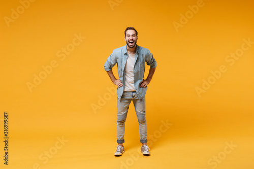 Laughing young bearded man in casual blue shirt posing isolated on yellow orange background, studio portrait. People emotions lifestyle concept. Mock up copy space. Standing with arms akimbo on waist.