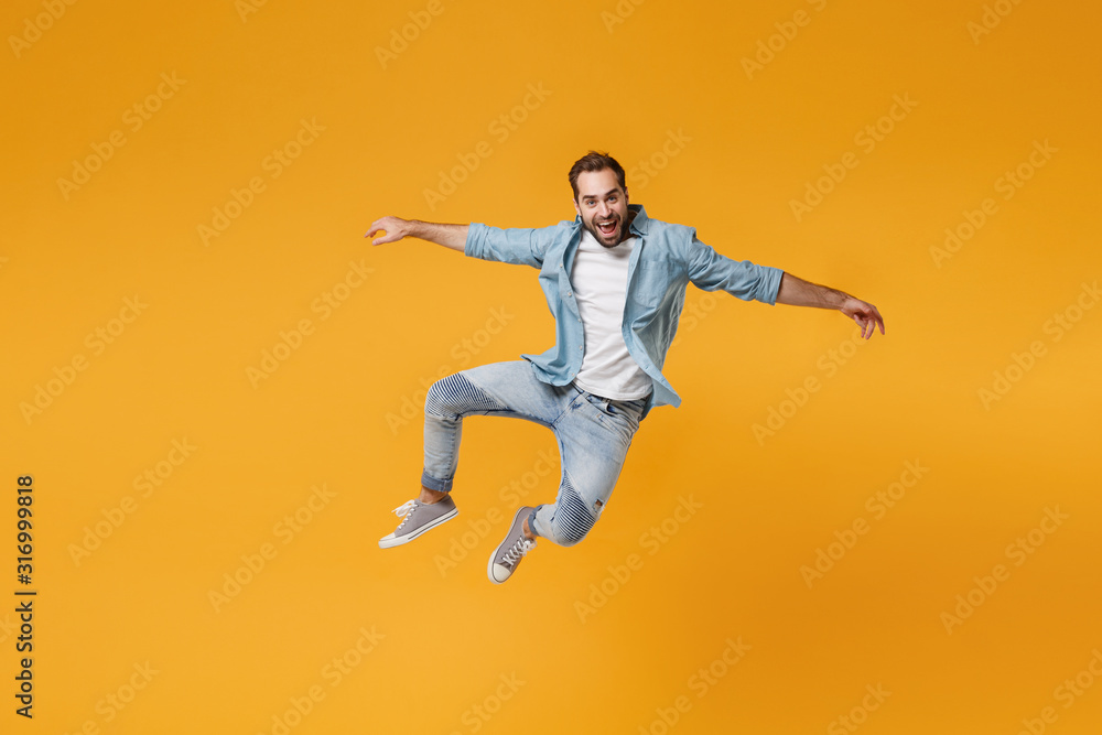 Cheerful young bearded man in casual blue shirt posing isolated on yellow orange background, studio portrait. People sincere emotions lifestyle concept. Mock up copy space. Jumping spreading hands.