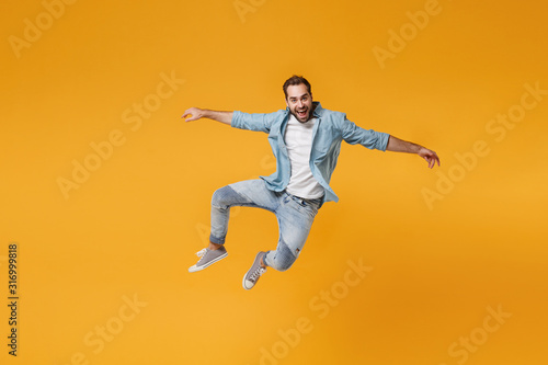 Cheerful young bearded man in casual blue shirt posing isolated on yellow orange background, studio portrait. People sincere emotions lifestyle concept. Mock up copy space. Jumping spreading hands.