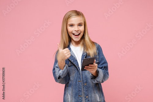 Funny little blonde kid girl 12-13 years old in denim jacket posing isolated on pastel pink background in studio. Childhood lifestyle concept. Mock up copy space. Hold mobile phone, showing thumb up.