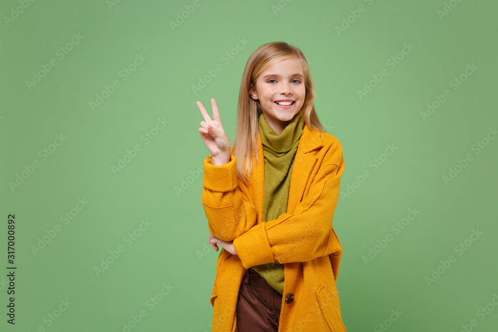 Pretty little blonde kid girl 12-13 years old in yellow coat posing isolated on pastel green background children studio portrait. Childhood lifestyle concept. Mock up copy space. Showing victory sign. Pretty little blonde kid girl 12-13 years old in yellow coat posing isolated on pastel green background children studio portrait. Childhood lifestyle concept. Mock up copy space. Showing victory sign.