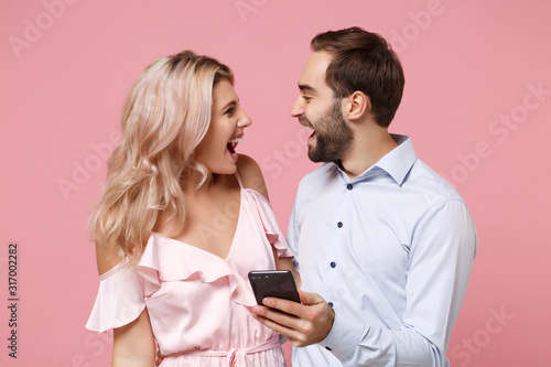 Cheerful young couple two guy girl in party outfit celebrating isolated on pastel pink background. Valentine's Day Women's Day birthday holiday party concept. Using mobile phone, typing sms message.