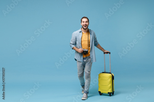 Laughing traveler tourist man in yellow casual clothes with photo camera, suitcase isolated on blue background. Male passenger traveling abroad on weekends. Air flight journey concept. Looking camera. photo