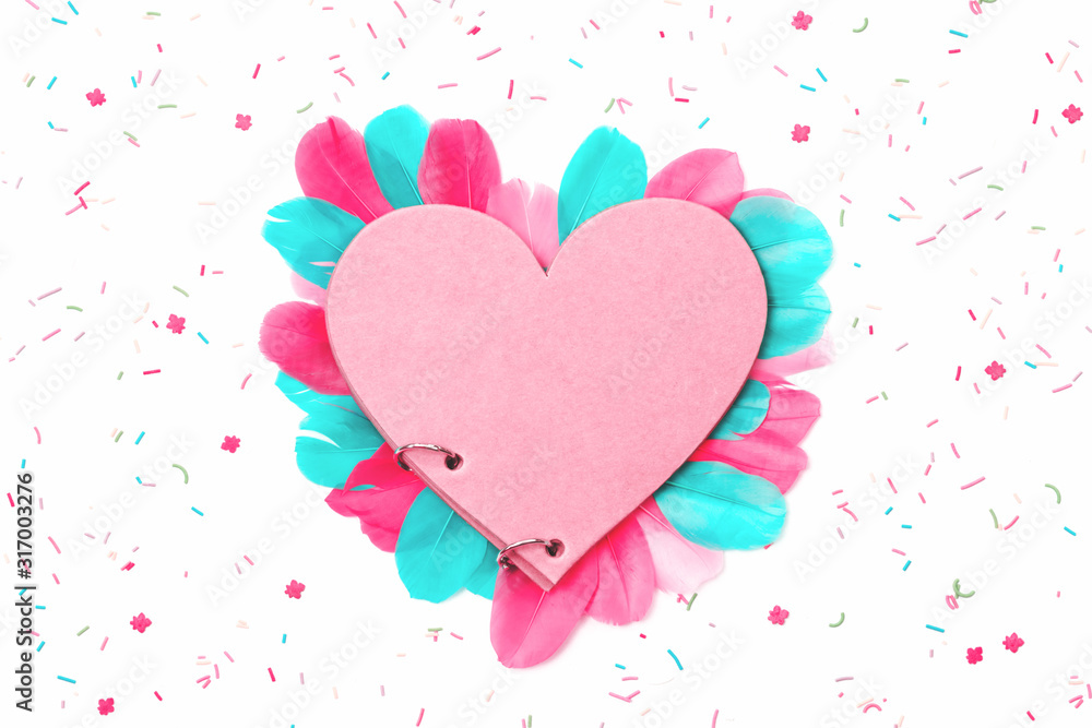 Creative love heart notepad with pink and mint feathers and scattered confetti and cake sprinkles isolated on white bavkground. St Valentines or Mothers Day, anniversary concept. Flat lay copy space.