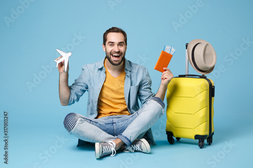 Cheerful traveler tourist man in yellow clothes isolated on blue background. Passenger traveling abroad on weekend. Air flight journey Sit near suitcase hold passport boarding pass tickets air plane. photo