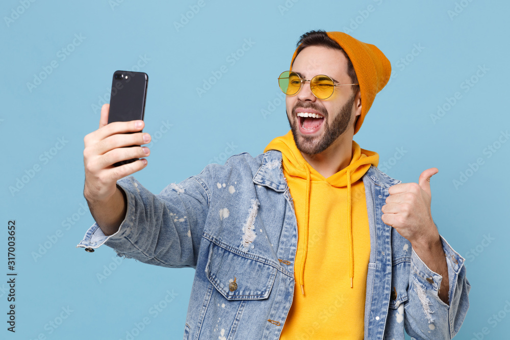 © ViDi Studio - Funny young hipster guy in fashion jeans denim clothes isolated on pastel blue background. People lifestyle concept. Mock up copy space. Doing selfie shot on mobile phone, showing thumb up, blinking.