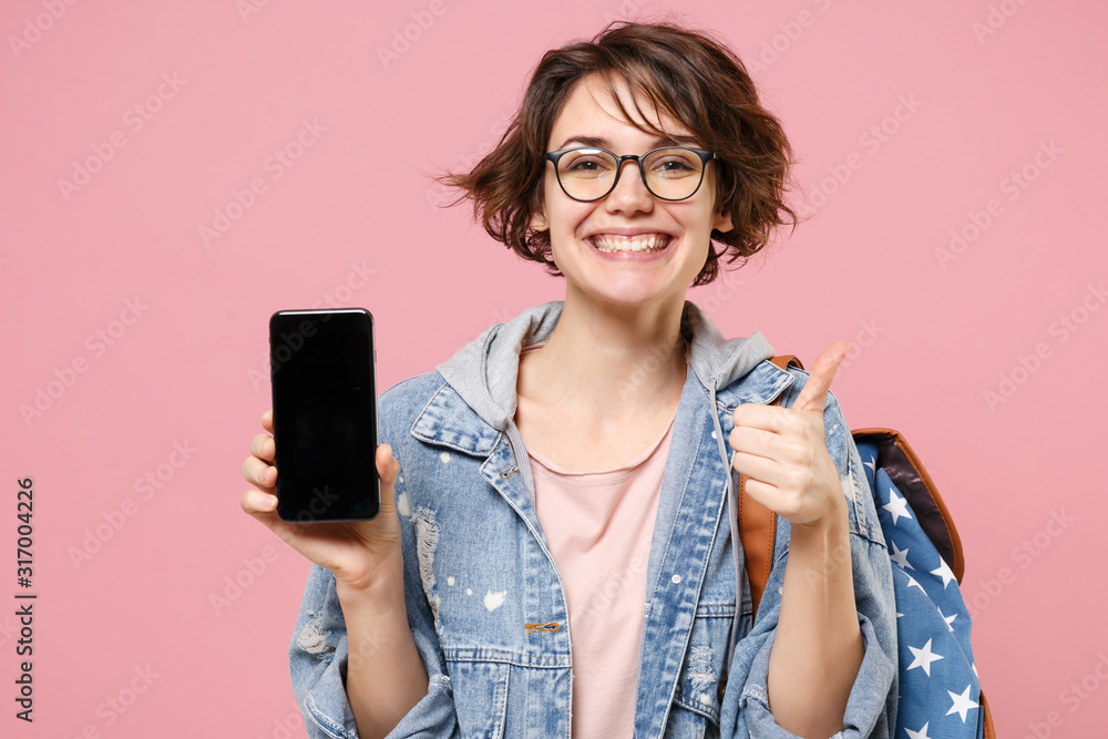 Smiling woman student in denim clothes glasses backpack isolated on pastel pink background. Education in school university college concept. Hold mobile phone with blank empty screen, showing thumb up. Smiling woman student in denim clothes glasses backpack isolated on pastel pink background. Education in school university college concept. Hold mobile phone with blank empty screen, showing thumb up.