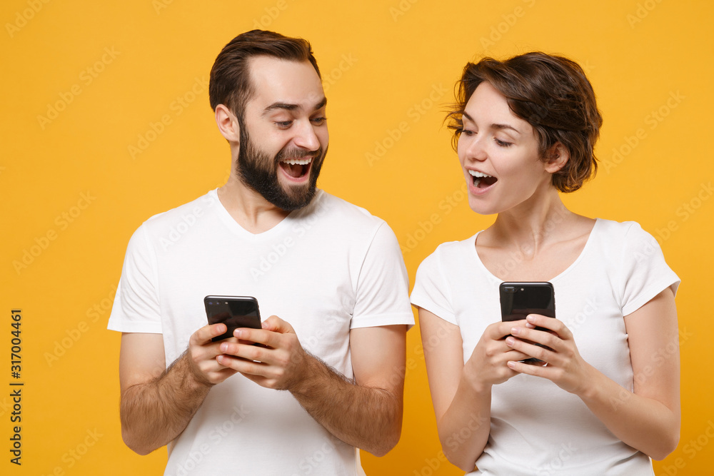 Excited young couple friends bearded guy girl in white blank empty t-shirts isolated on yellow orange background. People lifestyle concept. Mock up copy space. Using mobile phone, typing sms message. Excited young couple friends bearded guy girl in white blank empty t-shirts isolated on yellow orange background. People lifestyle concept. Mock up copy space. Using mobile phone, typing sms message.