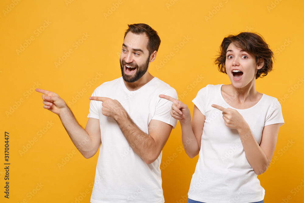 Surprised young couple friends bearded guy girl in white blank empty t-shirts posing isolated on yellow orange background. People lifestyle concept. Mock up copy space. Pointing index fingers aside. Surprised young couple friends bearded guy girl in white blank empty t-shirts posing isolated on yellow orange background. People lifestyle concept. Mock up copy space. Pointing index fingers aside.