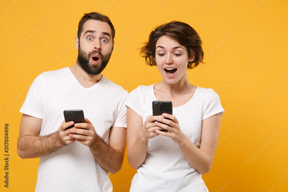 Amazed young couple friends bearded guy girl in white blank empty t-shirts isolated on yellow orange background. People lifestyle concept. Mock up copy space. Using mobile phone, typing sms message. Amazed young couple friends bearded guy girl in white blank empty t-shirts isolated on yellow orange background. People lifestyle concept. Mock up copy space. Using mobile phone, typing sms message.