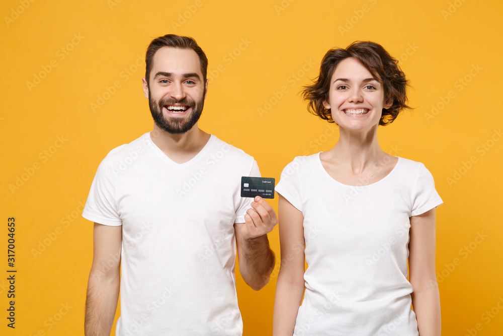 Smiling young couple friends bearded guy girl in white blank empty t-shirts isolated on yellow orange wall background in studio. People lifestyle concept. Mock up copy space. Holding credit bank card. Smiling young couple friends bearded guy girl in white blank empty t-shirts isolated on yellow orange wall background in studio. People lifestyle concept. Mock up copy space. Holding credit bank card.
