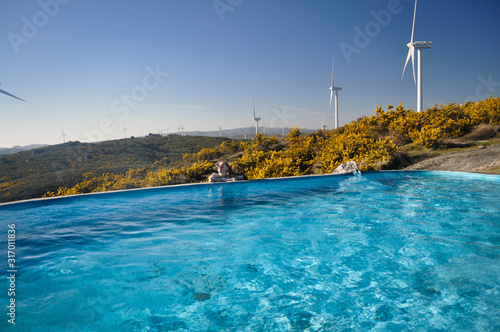 Blue water in the pool in the background wind farm behind the field yellow bushes . The Flintstone House Portugal