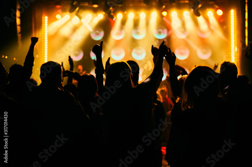 Crowd at a music concert, audience raising hands up in front of bright stage lights.