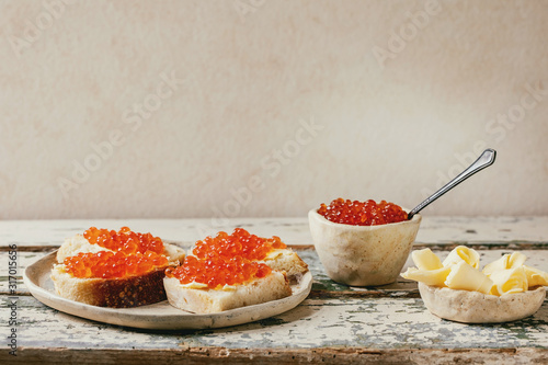 Red salmon caviar in bowl and on wheat bread, served with butter on ceramic plate over old wooden table. Copy space