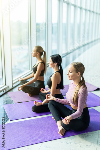 Three young women meditating in lotus pose during yoga class in health club. Women sitting on floor near window with city view