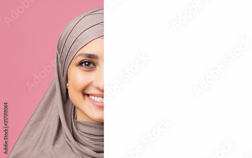 Smiling arabic woman in hijab standing behind white advertisement board © Prostock-studio