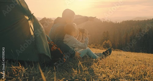 Happy traveler father and son sitting on grass and look through binoculars, bonding around mountain under sun light enjoying the leisure and freedom. 4K slow motion video