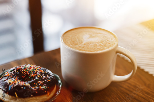 Coffee with a drawn heart and milk on a wooden table in a coffee shop. Chocolate donut with scattering on the table next to the coffee photo