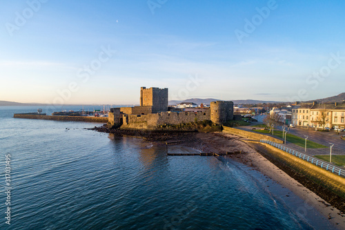Medieval Norman Castle in Carrickfergus near Belfast in sunrise light. Aerial view with marina, yachts,  breakwater, groyne, sediments and far view of Belfast in the background