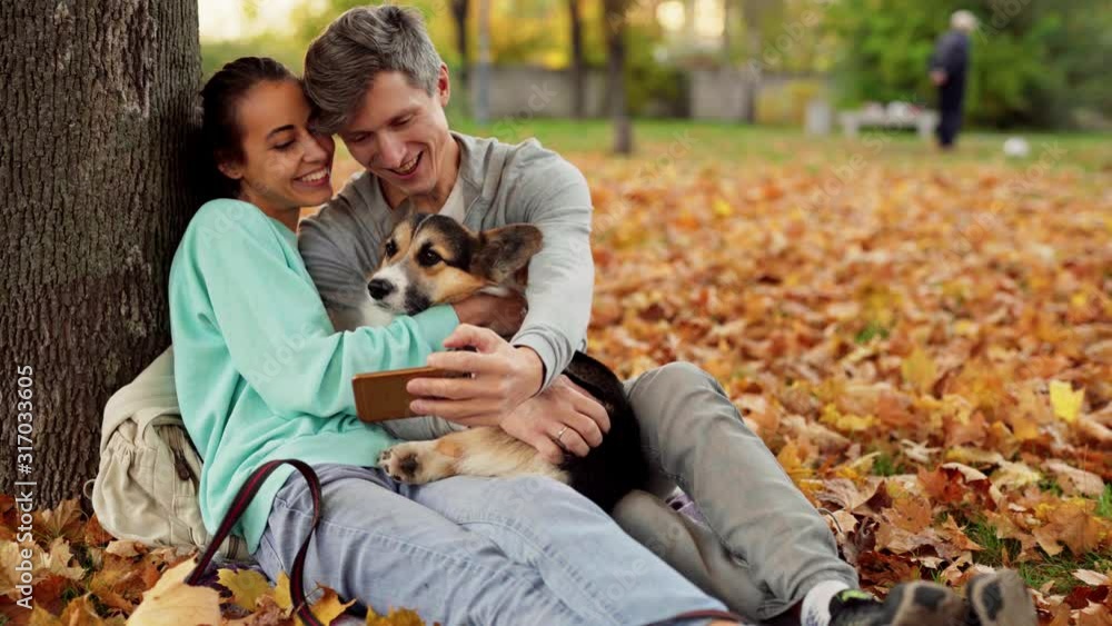 Loving Couple Man And Woman sitting with cute tricolor Welsh Corgi Dog In City Park hugging relaxing enjoying warm day outdoors and making selfie on smartphone. People and animals concept. Loving Couple Man And Woman sitting with cute tricolor Welsh Corgi Dog In City Park hugging relaxing enjoying warm day outdoors and making selfie on smartphone. People and animals concept.